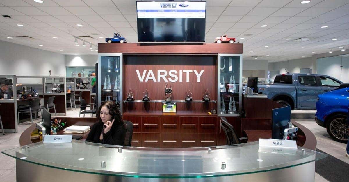 The reception area of Varsity Ford featuring a curved glass-top desk with nameplates for Emma and Alisha, a large wooden display with "VARSITY" branding, trophies, and a screen showing customer service details. The background includes office cubicles and display vehicles in a well-lit dealership setting.