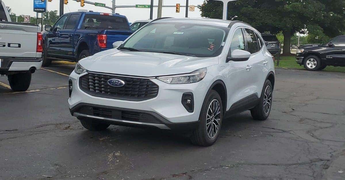 A white 2026 Ford Escape is parked diagonally at Varsity Ford, featuring a distinctive front grille with the Ford logo. Surrounding vehicles include a white and blue pickup truck. The background shows a 'Certified Pre-Owned Suburban' sign, street signs, and traffic lights, with trees and an overcast sky.
