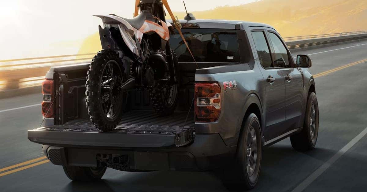 A metallic grey 2026 Ford Maverick FX4 pickup truck driving on a highway with a white and orange dirt bike secured in its open tailgate. The truck features dark alloy wheels and "FX4 OFF ROAD" badging. The background includes rolling hills and a hazy sky, emphasizing adventure and utility.