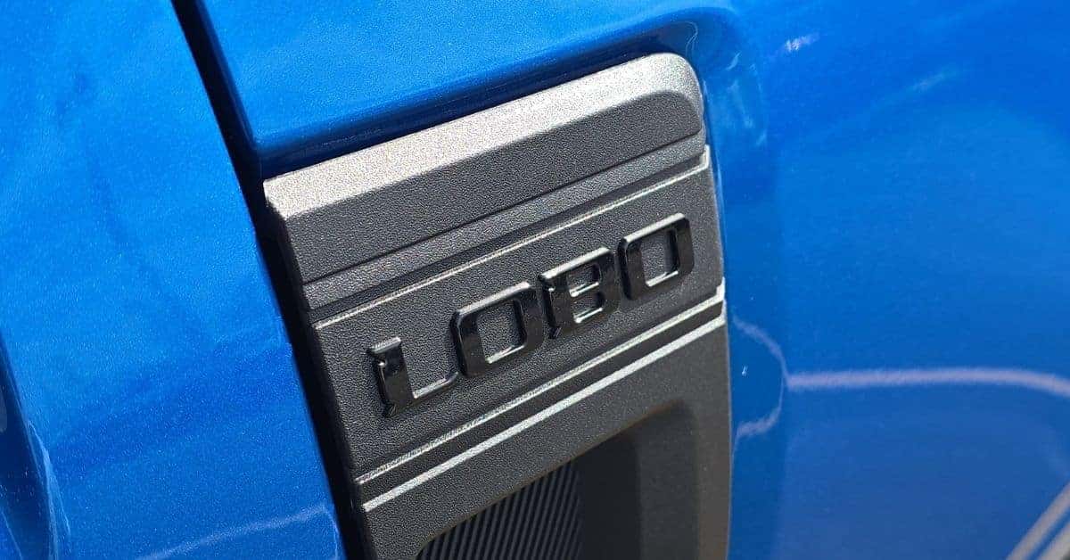 A close-up of the "LOBO" fender badge on a bright blue 2026 Ford Maverick truck. The badge features glossy black embossed letters on a textured grey trim piece, set against the metallic blue body panel. A vent with a ribbed texture is visible below the badge, showcasing modern automotive styling.