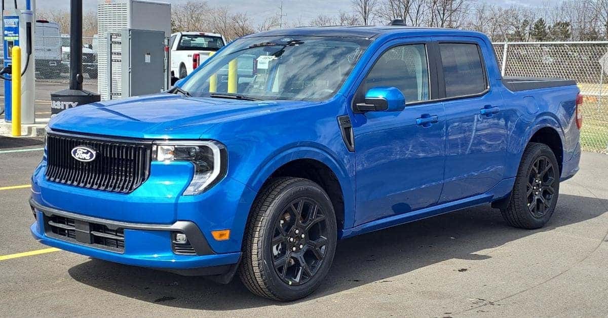 A bright blue 2026 Ford Maverick Lobo pickup truck parked in the Varsity Ford lot. The truck features a black grille, multi-spoke alloy wheels, and black trim accents. The background includes an EV charging station, a white pickup truck, and bare trees under a bright sky.