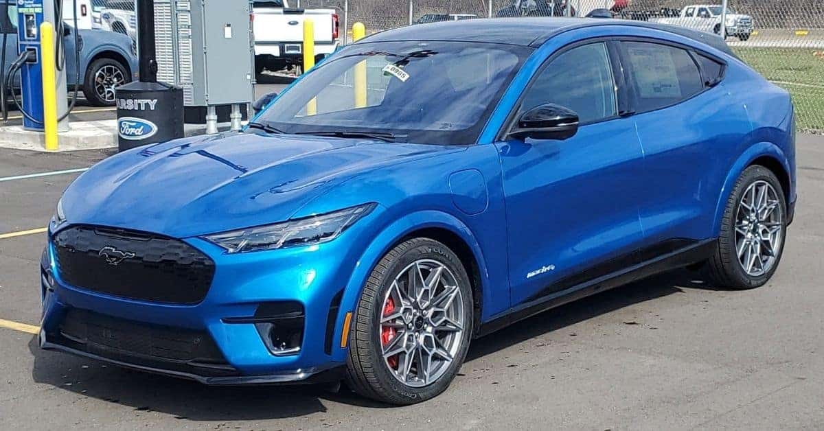 A vibrant blue 2026 Ford Mustang Mach-E GT parked in a dealership lot next to an EV charging station. The car features a black grille with a silver pony emblem, red Brembo brake calipers, and a panoramic black glass roof. The background includes a white Ford F-150 and Varisty Ford dealership branding.