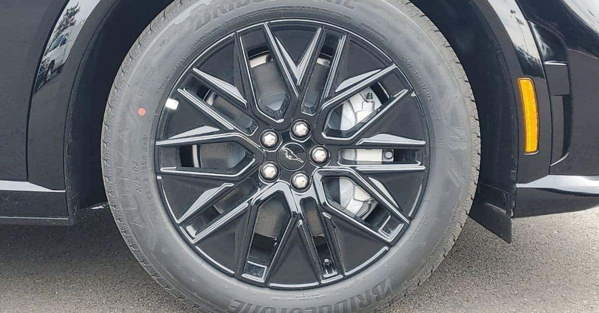A detailed close-up of a 19-inch Bridgestone Alenza tire mounted on a black and silver machined alloy wheel of a 2026 Ford Mustang Mach-E. The wheel features a Mustang pony logo on the center cap, with a glossy black fender and amber side marker reflector visible.