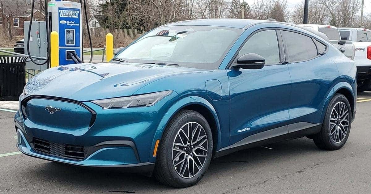 A teal 2026 Ford Mustang Mach-E electric SUV parked at a Varsity Ford-branded ChargePoint charging station. The car features a sleek design with a closed front grille, multi-spoke alloy wheels, and a visible charging port on the front fender. The background includes a brick building, bare trees, and parked vehicles, emphasizing the integration of EV technology with modern infrastructure.