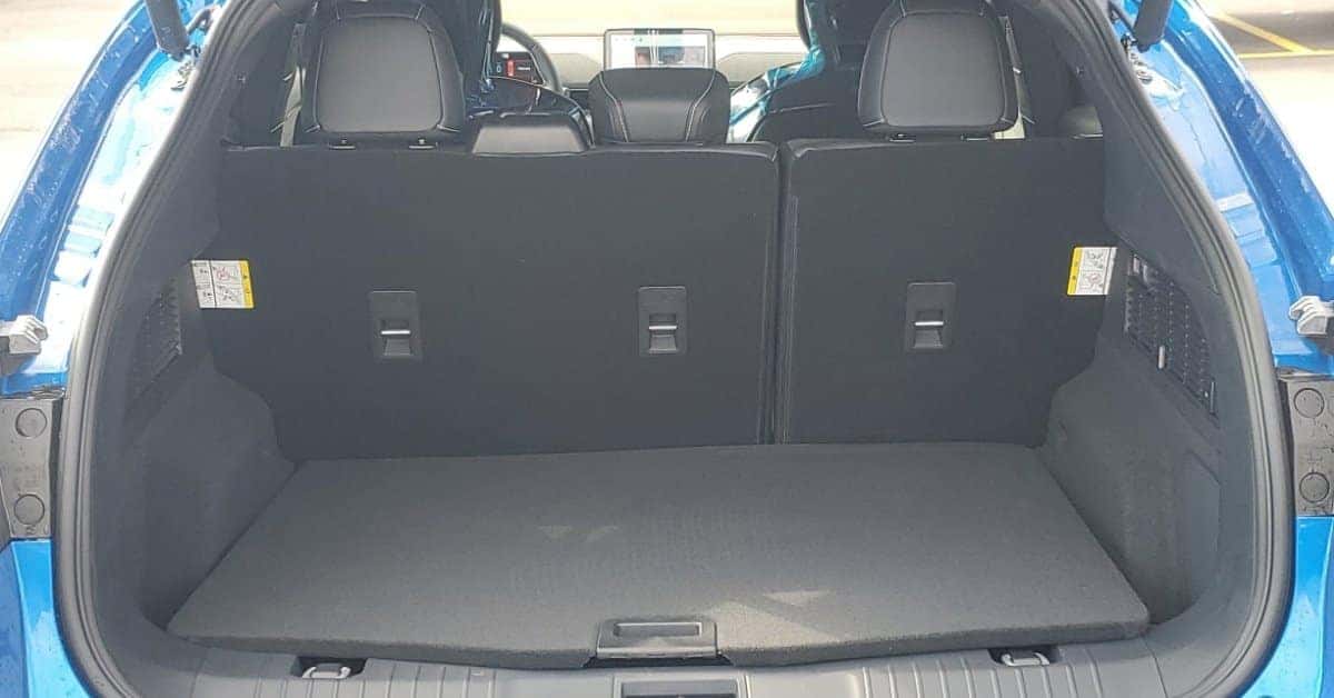 The rear cargo area of a bright blue 2026 Ford Mustang Mach-E with the liftgate open, showcasing split-folding rear seats with tether anchors, a flat carpeted trunk floor, and protective plastic on the front seats. The image highlights the vehicle's spacious interior and practical design, suitable for showcasing cargo capacity and modern features.