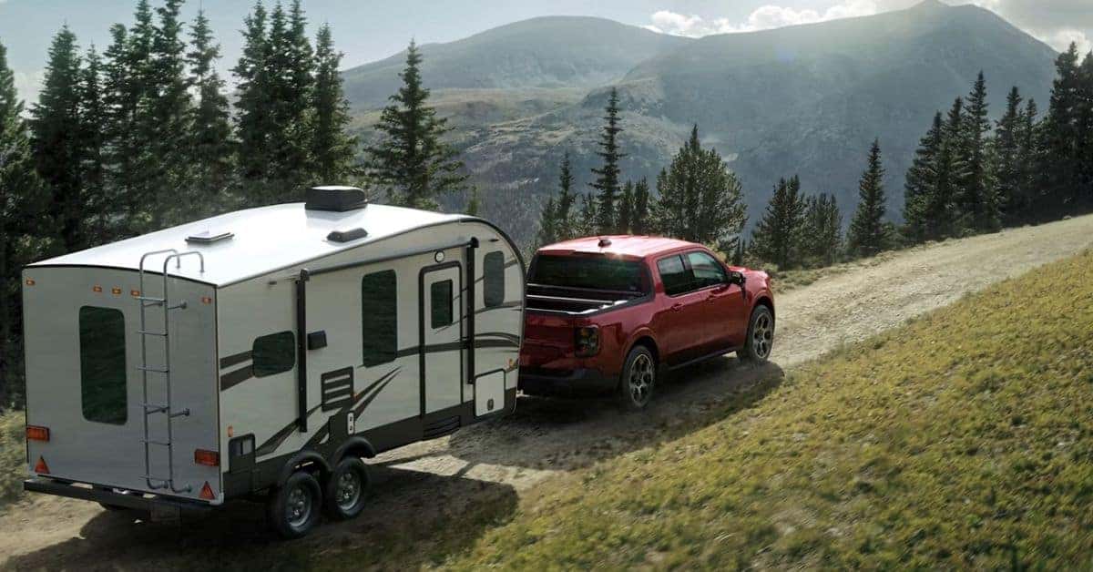 A red four-door 2026 Ford Maverick pickup truck towing a white dual-axle travel trailer up a dirt mountain road. The scene features green grassy hills, dense evergreen trees, and towering blue mountains in the background, highlighting the truck's towing capability and rugged performance.