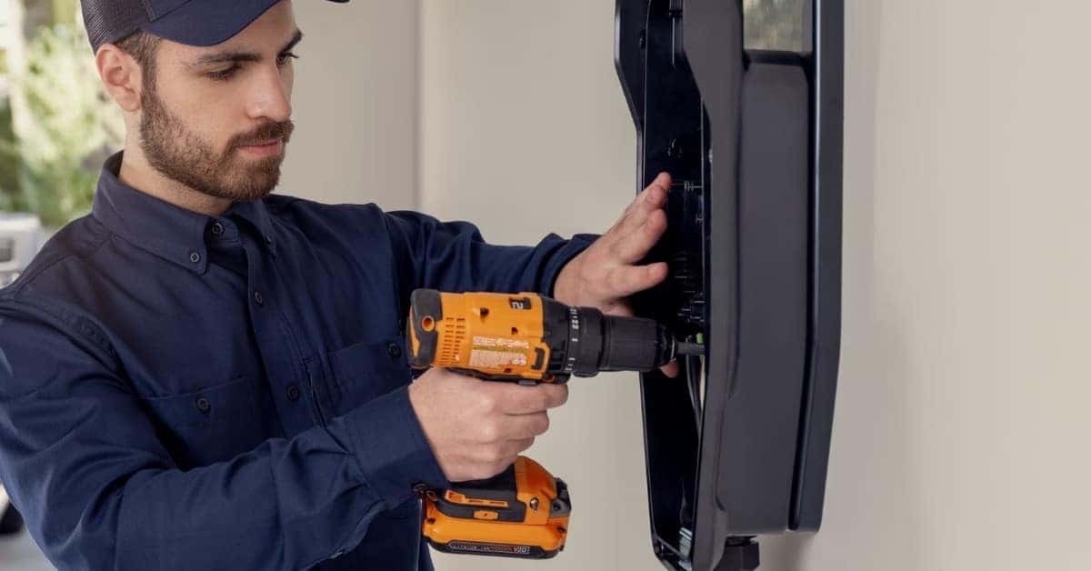A male technician in a navy uniform uses a black and orange cordless drill to install or repair a black wall-mounted EV charging station. The scene is set in a clean garage or carport with green foliage and a white vehicle visible in the blurred background.