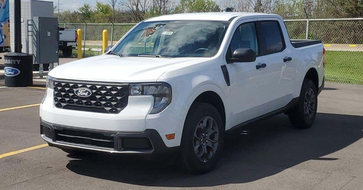 A white 2026 Ford Maverick crew cab pickup truck parked at the Varsity Ford dealership. The truck features a black mesh grille with a silver Ford emblem, C-shaped LED headlights, black trim accents, and dark grey multi-spoke alloy wheels. The background includes a branded black bin, a chain-link fence, green trees, and a blue sky with light clouds.
