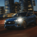2024 Ford Mustang Mach-E driving with its headlights on with a cityscape in the background