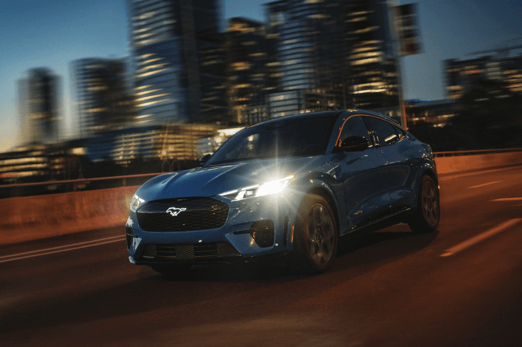 2024 Ford Mustang Mach-E driving with its headlights on with a cityscape in the background