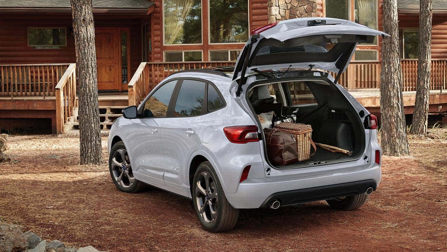 A silver Ford Escape Hybrid with its trunk open, parked in front of a log cabin