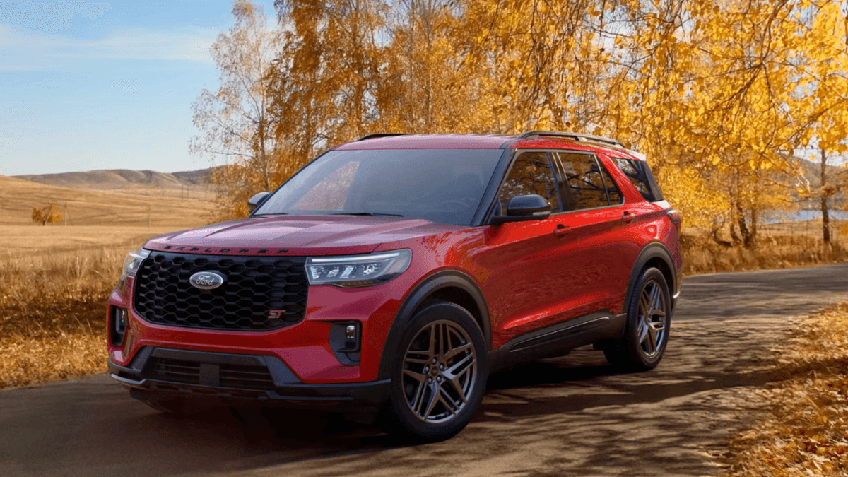 A red Ford Explorer driving down the road past a field and trees in autumn