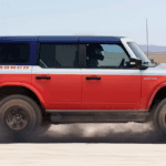 A red and white Ford Bronco in profile driving over a desert trail and kicking up dust
