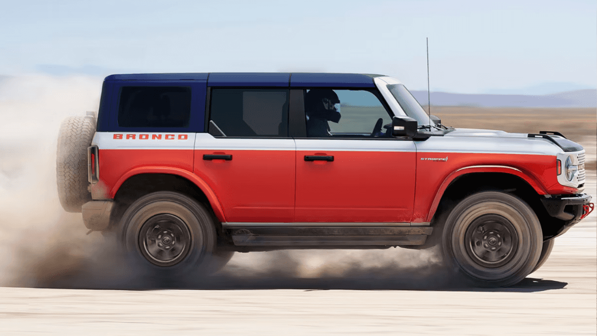 A red and white Ford Bronco in profile driving over a desert trail and kicking up dust