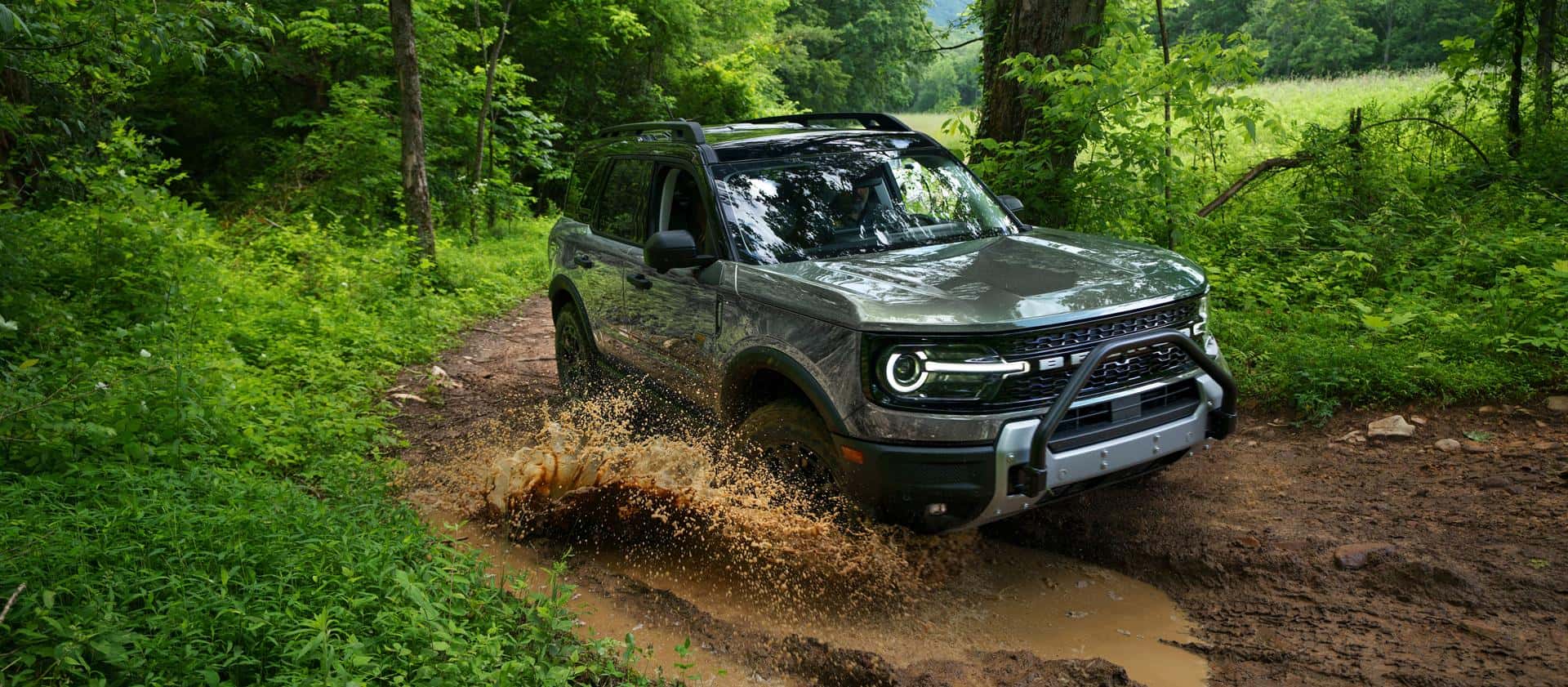 A light green Ford Bronco Sport driving through a muddy dirt road in a forest