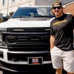 Man wearing black tee shirt and white shorts standing in front of white Ford truck