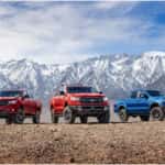 Two red Ford Rangers and a blue one parked on dirt with snow covered mountains in the background.
