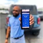 A hand holding a smartphone showing the FordPass app with a gray Ford Bronco in the background.