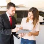 A man in a black jacket and a woman in a beige top holding a clipboard in front of a white car.