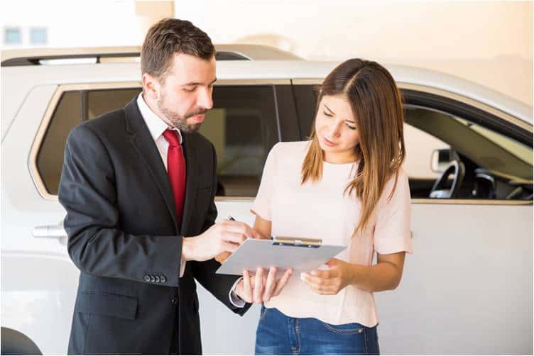 A man in a black jacket and a woman in a beige top holding a clipboard in front of a white car.