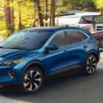 A blue 2025 Ford Escape towing a boat along a road on the water