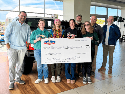 A group of students and adults standing inside a dealership showroom, holding a large novelty check for $2,070 made out to North Central High School.