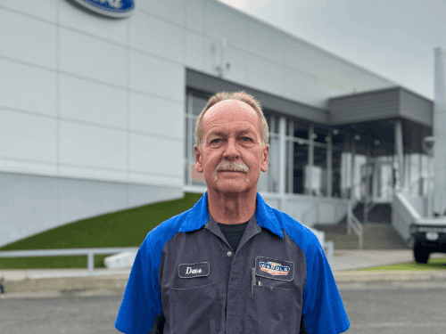 A man wearing a blue and gray uniform work shirt standing in front of the Wendle Ford dealership building.