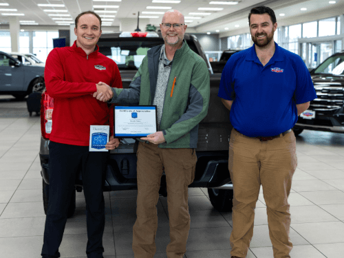 Three men standing in a the Wendle Ford showroom in front of a truck. Two men are shaking hands while holding a framed Certificate of Appreciation and a thank you card featuring the Spokane Police Foundation logo.