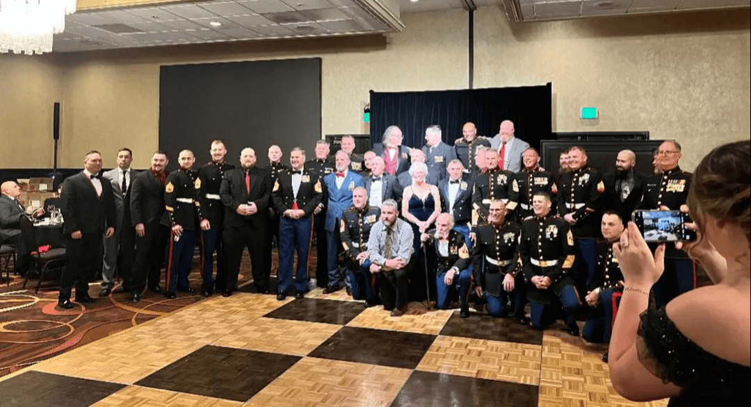 A large group of US Marines in dress blues and guests posing for a photo at the 250th US Marine Corps Birthday Ball.