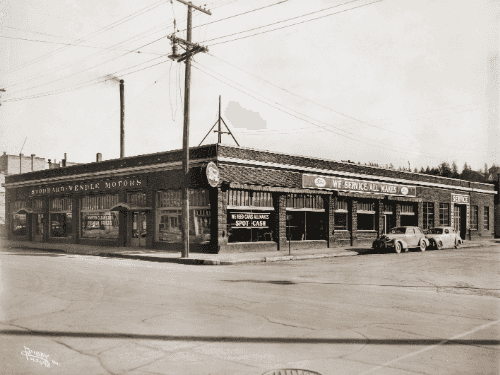 Exterior view of Stoddard-Wendle Motors in 1944, a Ford dealership in Spokane WA. The brick building features signs for 'Service,' 'We Service All Makes,' and 'We Need Cars All Makes,' with vintage 1940s vehicles parked out front.