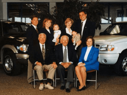 A multi-generational portrait of the Wendle family posing together in a vehicle showroom between a dark SUV and a white truck.
