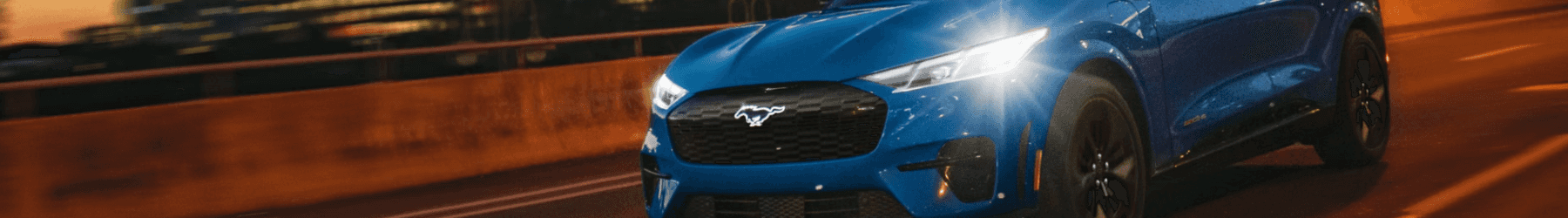 A bright blue Ford Mustang Mach-E electric SUV driving on a city highway at night with blurred skyscraper lights in the background.