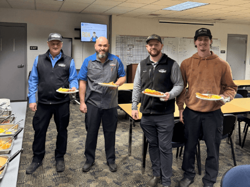 Four Wendle Ford service technicians smiling and holding plates of food at a staff appreciation breakfast. The team is gathered in the dealership breakroom for a community-building meal.