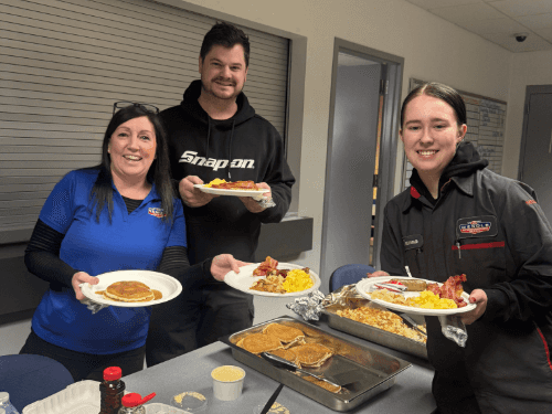 Three Wendle service staff smiling and holding plates of food at a staff appreciation breakfast. The team is gathered in the dealership breakroom for a community-building meal.