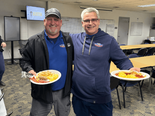 Two Wendle service staff smiling and holding plates of food at a staff appreciation breakfast. The team is gathered in the dealership breakroom for a community-building meal.