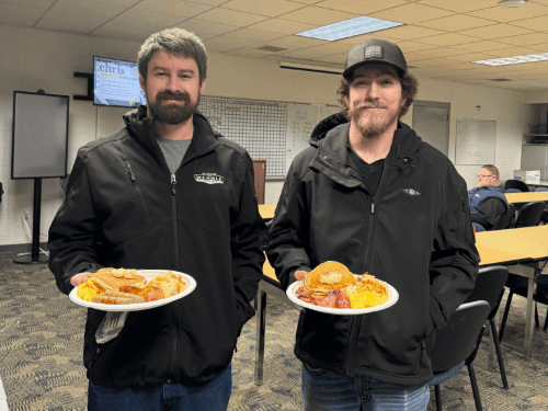 Two Wendle service staff smiling and holding plates of food at a staff appreciation breakfast. The team is gathered in the dealership breakroom for a community-building meal.