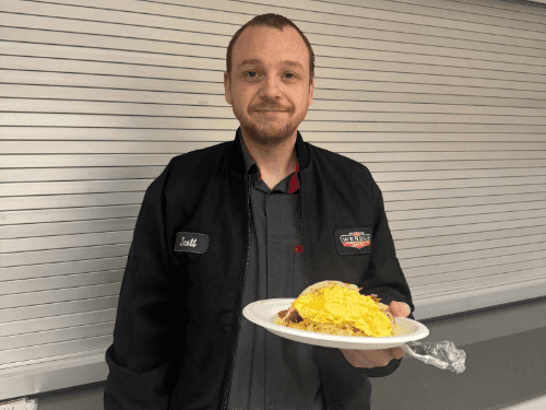 A Wendle service staff smiling and holding plates of food at a staff appreciation breakfast. The team is gathered in the dealership breakroom for a community-building meal.