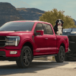 A red Ford F-150 SuperCrew with a dog in the bed is on a boat ramp, towing a black boat on a trailer, with hills and a lake in the background.