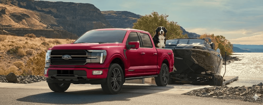 A red Ford F-150 SuperCrew with a dog in the bed is on a boat ramp, towing a black boat on a trailer, with hills and a lake in the background.