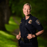 A smiling woman in a Spokane Police uniform standing outdoors with a blurred background of green trees.