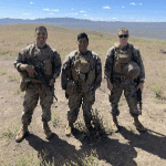 Three service members in military camouflage and tactical gear standing together in a dry, open field with hills in the background.