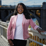 A woman smiling in a pink blazer and white turtleneck, leaning against a bridge railing with a river and city in the background.