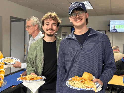 Two young men smile while holding plates of pasta and bread at a Wendle Ford event in a bright office breakroom setting.
