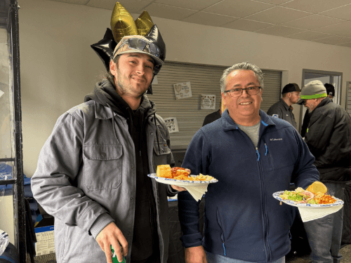 Two men holding food plates at a Wendle Ford celebration; one man wears a hat with gold and black balloons behind him.