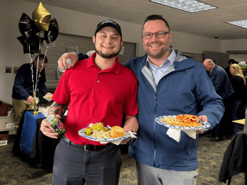 Two men smiling and holding plates of food at a Wendle Ford office celebration with gold and black balloons nearby.