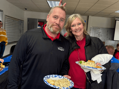 A man in a Wendle Ford pullover and a woman smile with plates of food; the woman playfully gives the man "bunny ears."