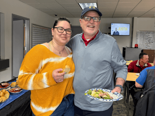 A woman in a striped sweater and a man in a grey pullover smile together with food plates at a Wendle Ford office event.