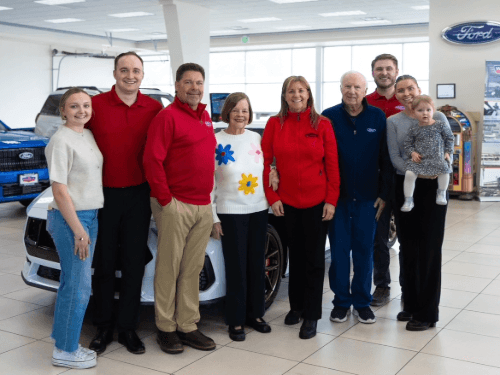 A large group of people and a small child smile while posing together in a bright Wendle Ford dealership showroom floor.