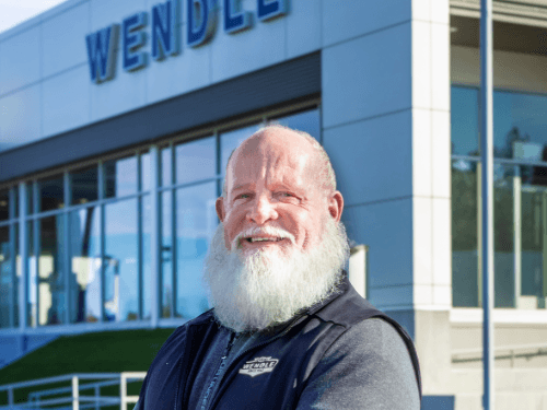 Smiling man with a white beard wearing a Wendle Ford vest stands in front of the modern Wendle dealership building.