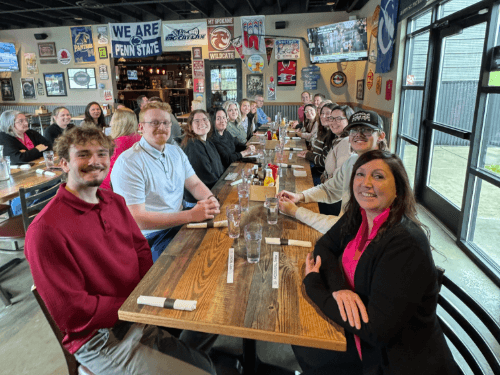 A large group of smiling dealership employees gathered around a long wooden table for a group lunch together at a restaurant.
